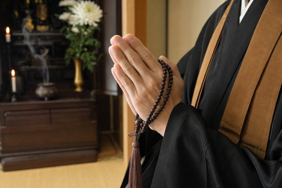 数珠を手に合掌する僧侶の手元と、仏壇（イメージ）